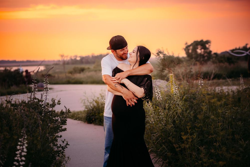 Two people share a tender kiss during sunset by a serene lake with tall grasses swaying in the breeze.
