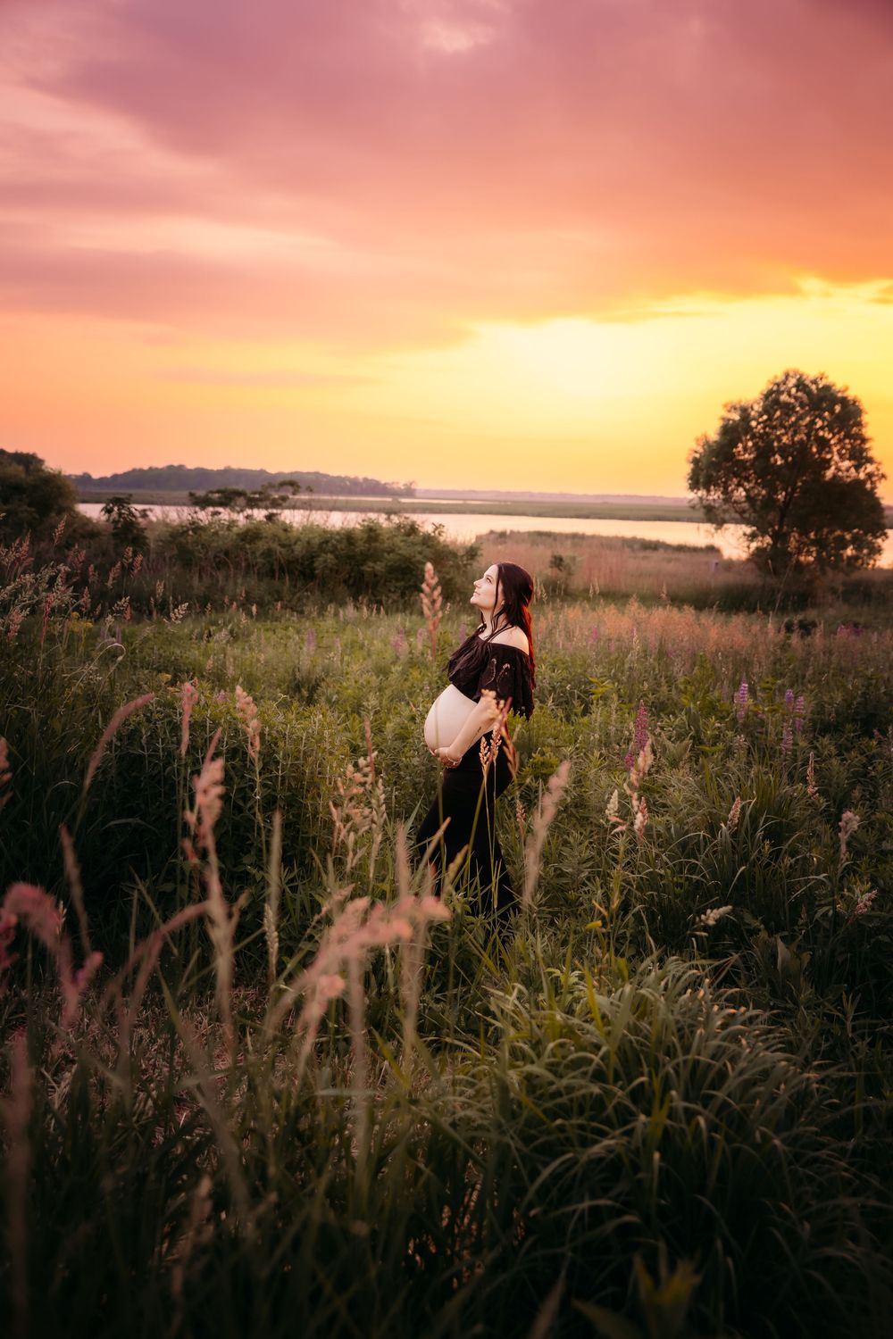 Silhouette against golden sunset sky stands among tall grass and wildflowers near tranquil waterfront at dusk.