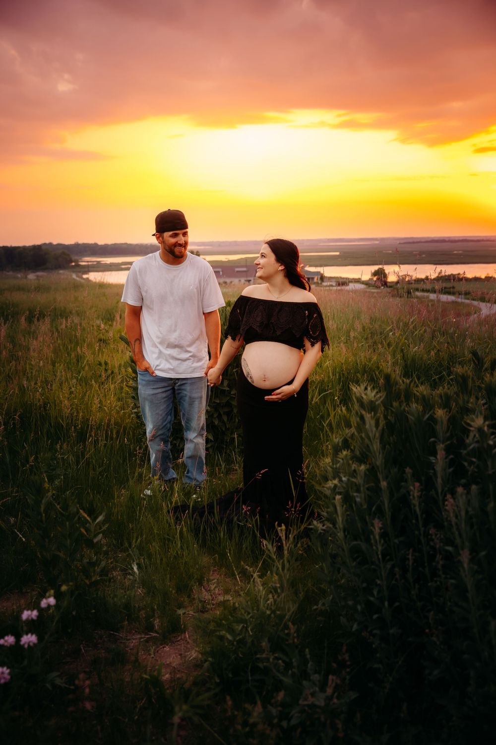 A couple stands in a grassy field at sunset, with the expectant mother wearing a black dress against the golden sky.