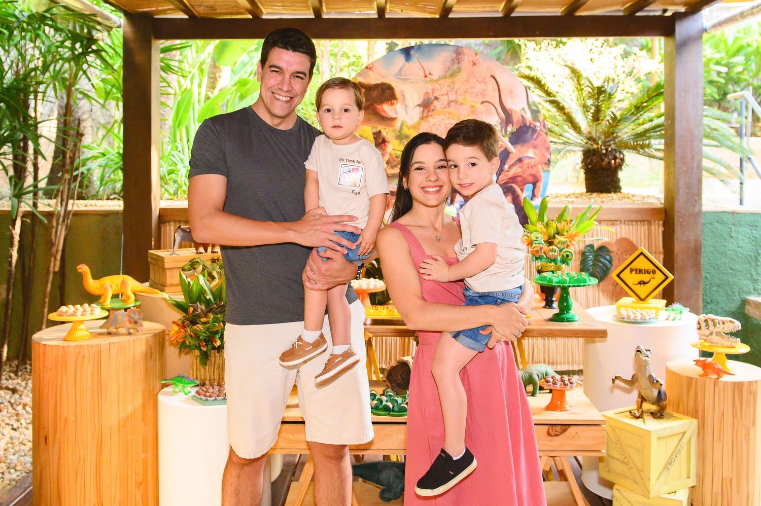 Family gathering around a decorated dessert table on an outdoor patio with wooden furniture and tropical plants.