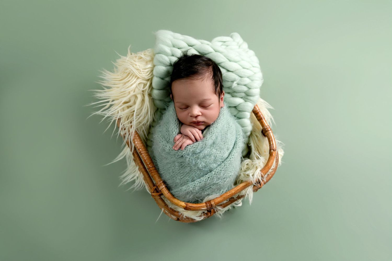 A sleeping newborn swaddled in mint green wrap rests peacefully in a round woven basket against mint background.