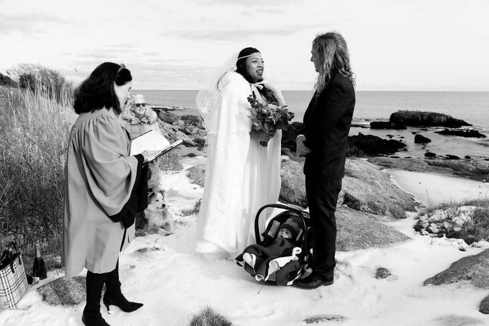 A winter beach wedding ceremony takes place on snowy rocks overlooking the ocean in black and white.
