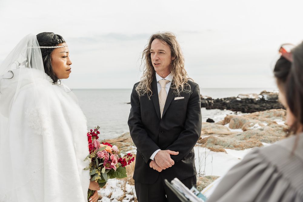 Two people in wedding attire stand on a rocky beach coastline during an outdoor ceremony.
