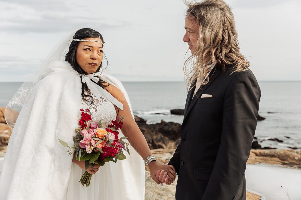 A couple holds hands during their seaside wedding ceremony with the bride in white holding a pink floral bouquet.