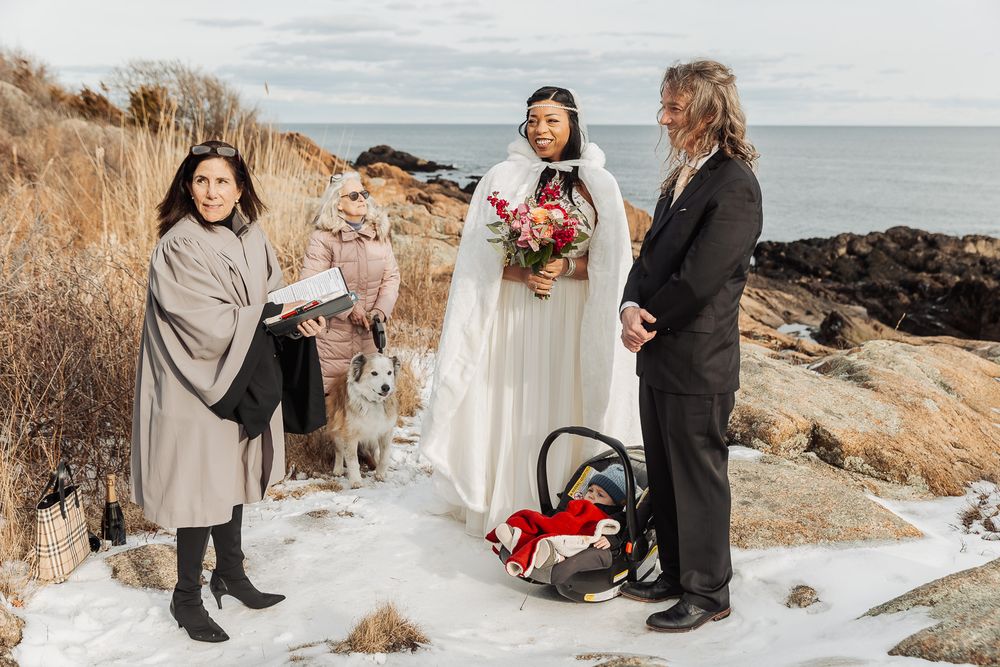A winter coastal wedding ceremony takes place on snow-covered rocky beach with tall grass and ocean views in background.
