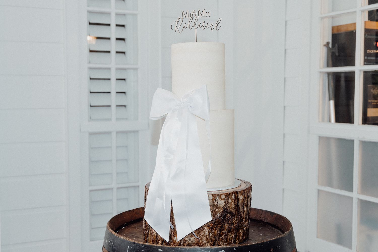 A tall white wedding cake with a large white bow sits on a rustic wood slice stand captured by Apertured Media Auckland Wedding Photographer