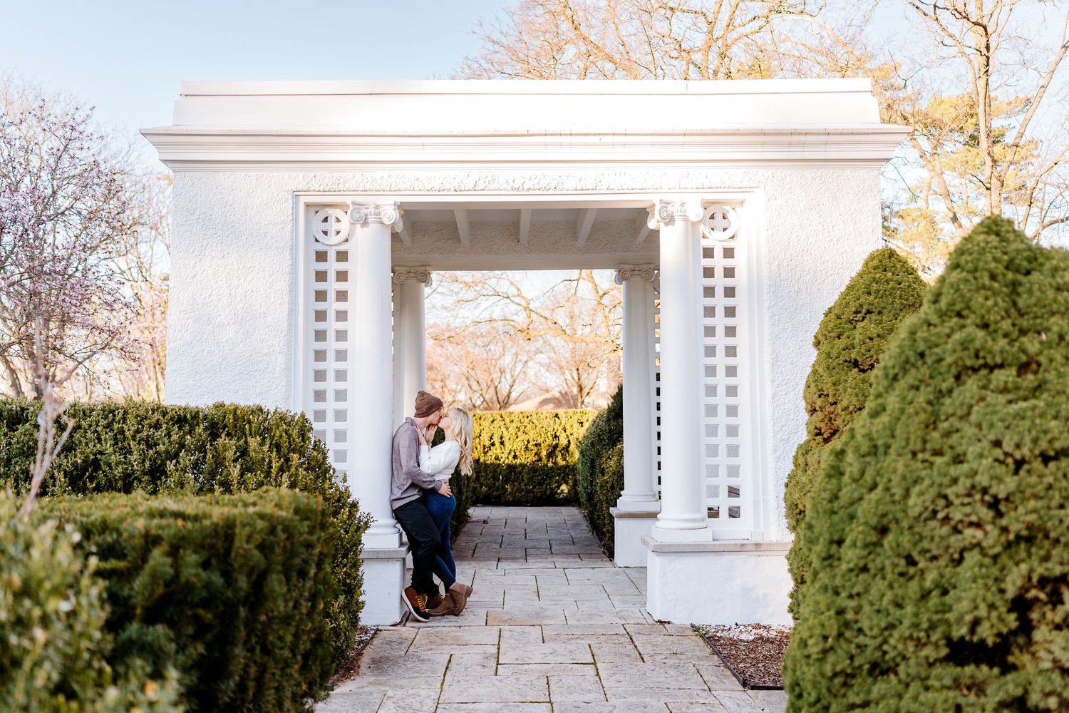 A white ornate garden archway with decorative columns stands between evergreen hedges on a stone pathway.