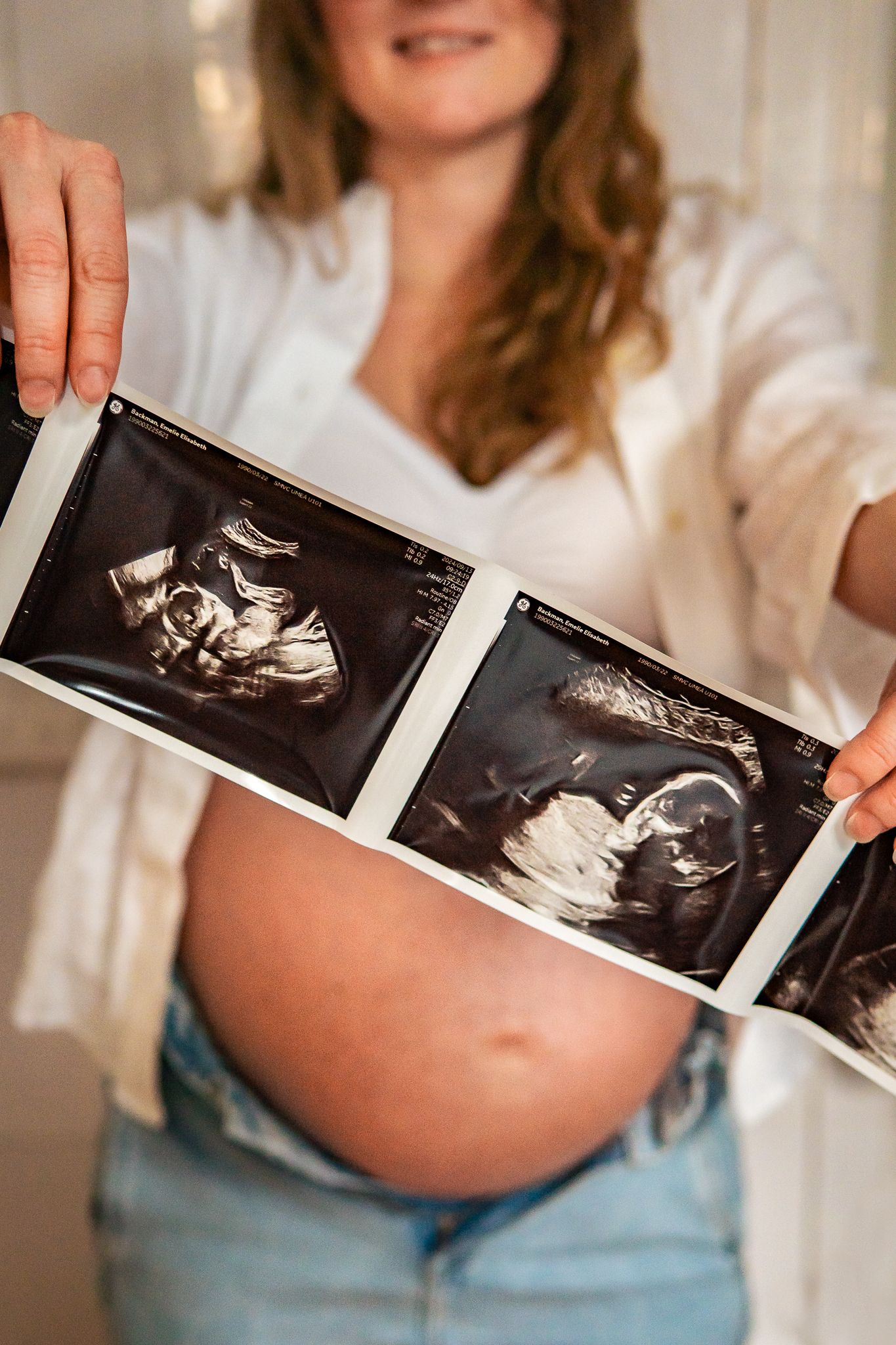 Gravid kvinna i vit bh och jeans håller ultraljudsbilder snett framför gravida magen. Gravidfotograf, Umeå, Västerbotten