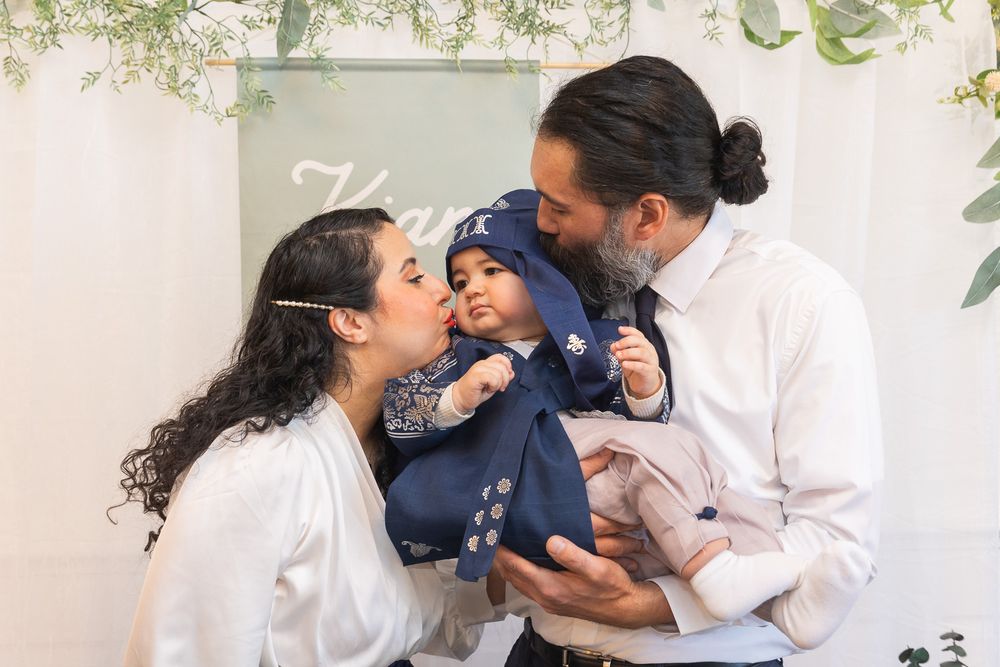 A young family shares a tender moment against a white backdrop with greenery overhead as they embrace their baby in navy blue.