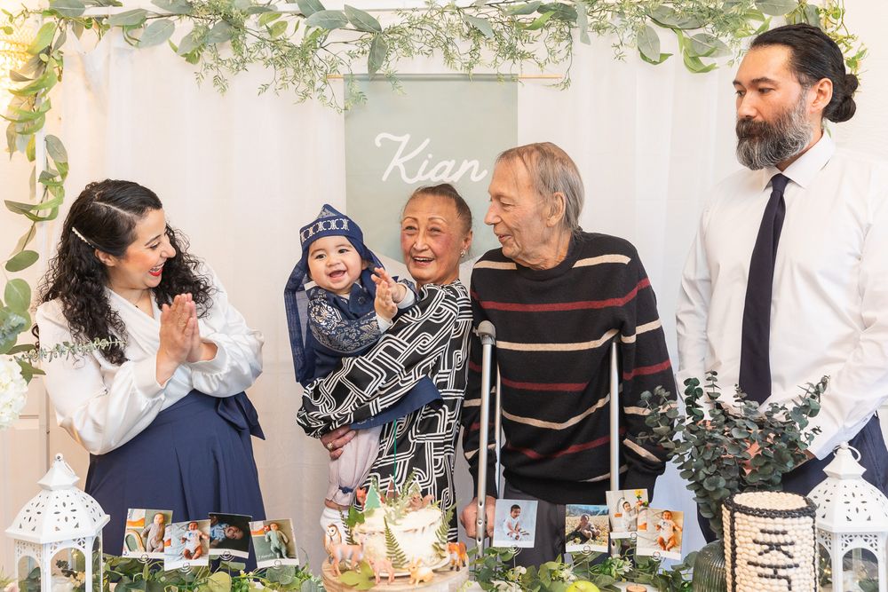 A group gathers around a decorated table with food and greenery for a special celebration under floral arch decor.