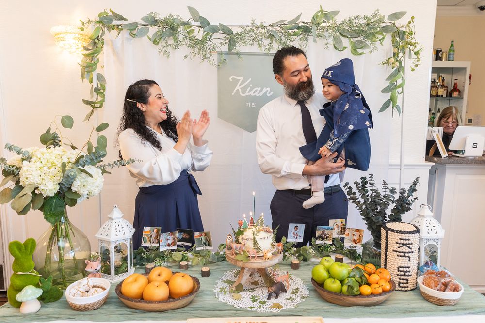 People celebrate around an elegantly decorated table with white flowers, fresh fruits and treats under a floral archway.