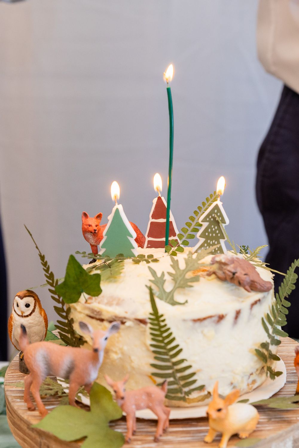 A whimsical woodland birthday cake decorated with green trees, candles, fern leaves and miniature forest animal figurines.