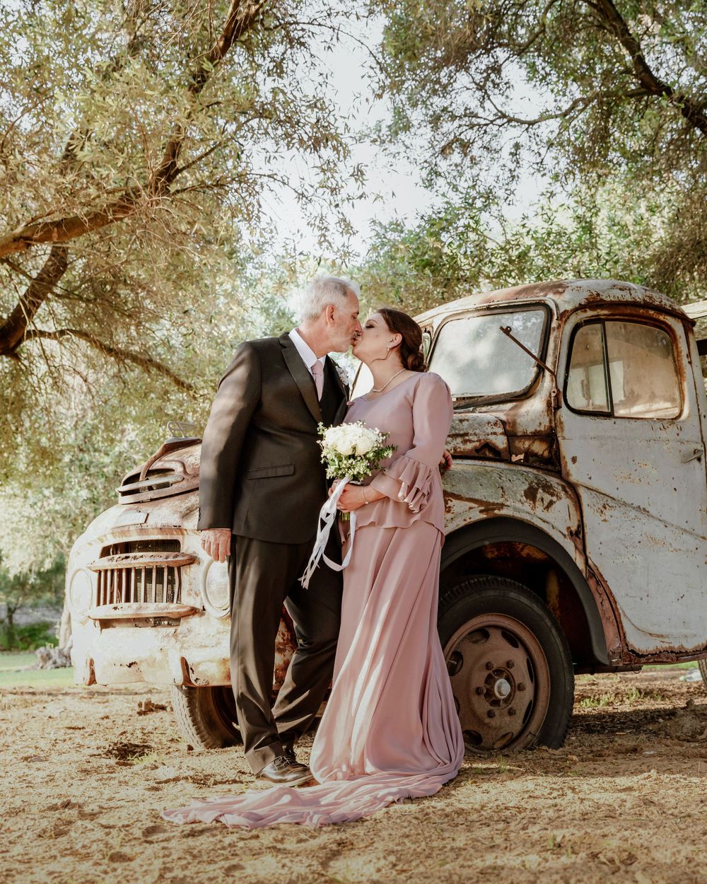 A romantic moment captured between newlyweds in front of an antique truck surrounded by trees.