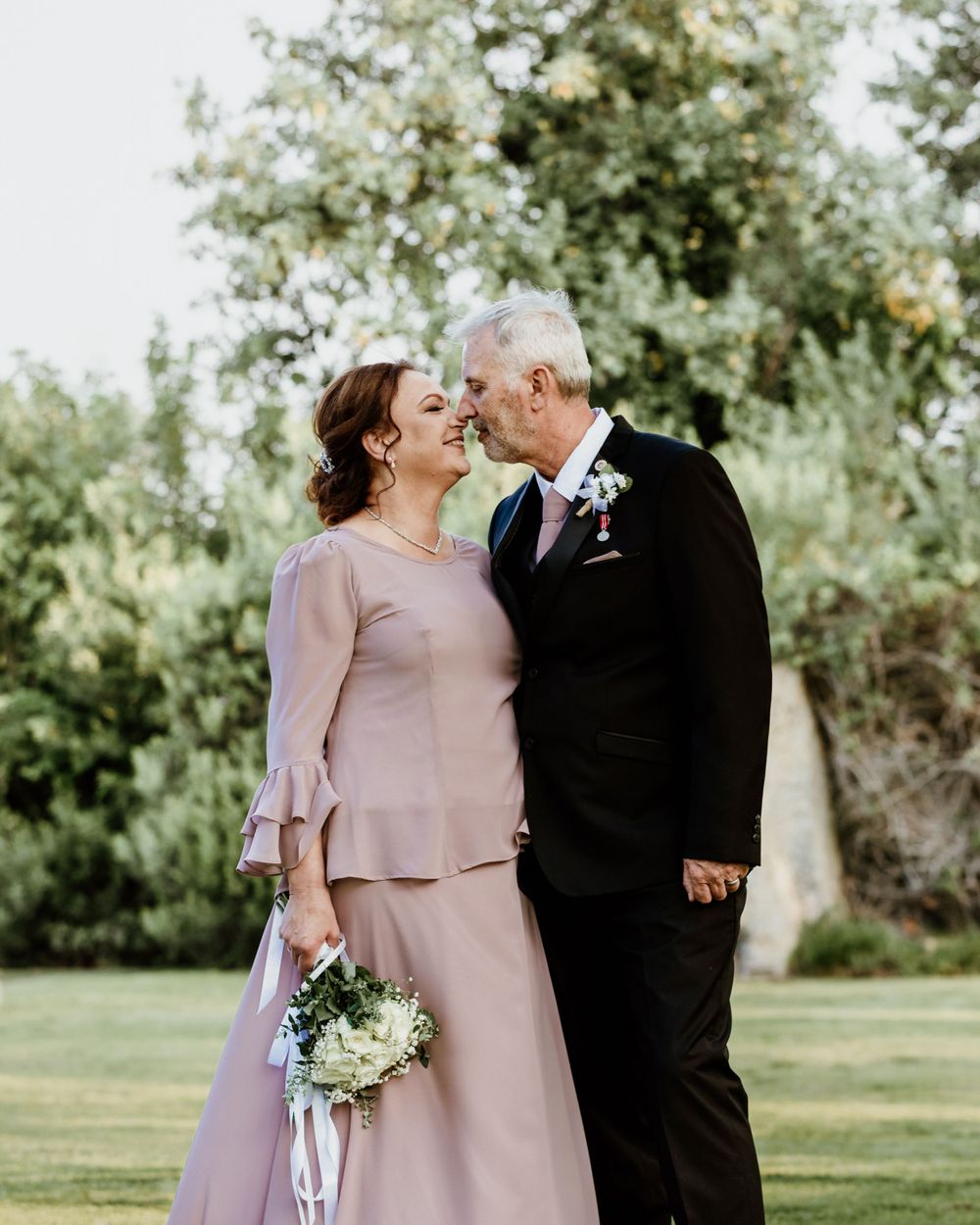 A tender outdoor scene in a garden with two people sharing a joyful embrace while one holds a delicate flower bouquet.
