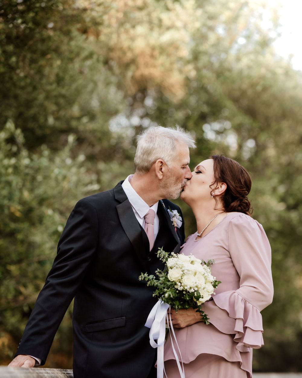 A couple shares a tender moment in a garden setting, wearing formal attire with a pink tie and mauve dress.