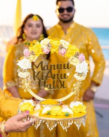 Yellow floral halo-style nameplate reading 'Mannu ki Anu Haldi' held up during a Haldi ceremony at the beach.