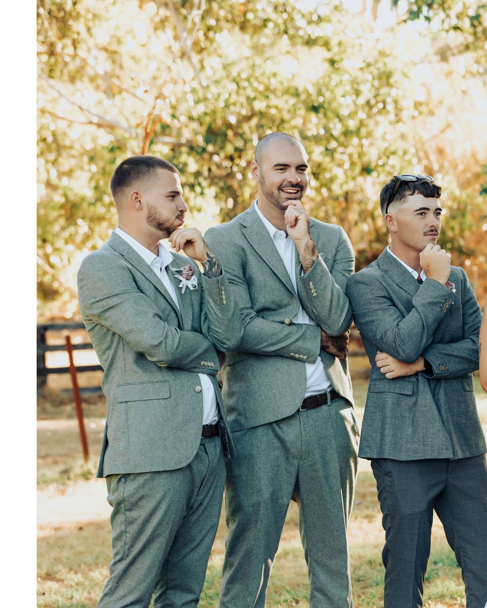 Three groomsmen in matching gray suits strike thoughtful poses while standing together at an outdoor wedding venue.