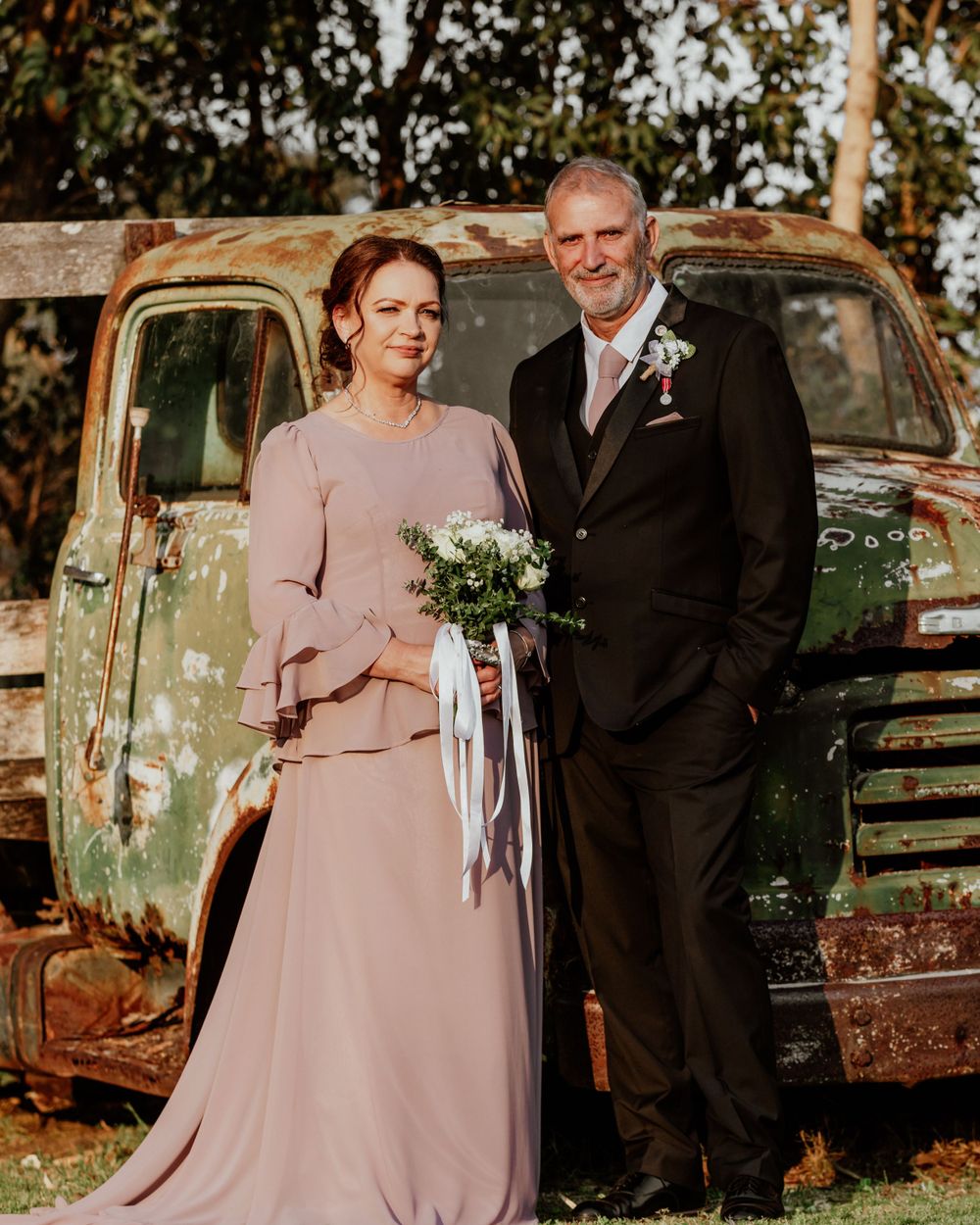 A couple poses in front of a rustic vintage truck, with the bride wearing a dusty pink gown and holding a bouquet.