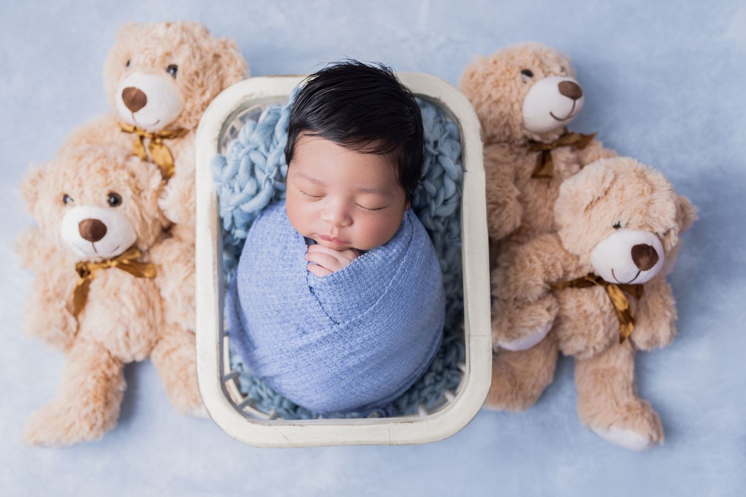 A sleeping newborn wrapped in blue swaddle blanket lies in white basket surrounded by soft beige teddy bears.