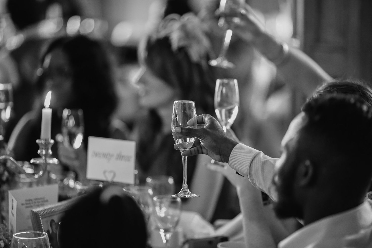 Wedding guests raise glasses during a celebratory toast at an elegant dinner reception table in black and white.