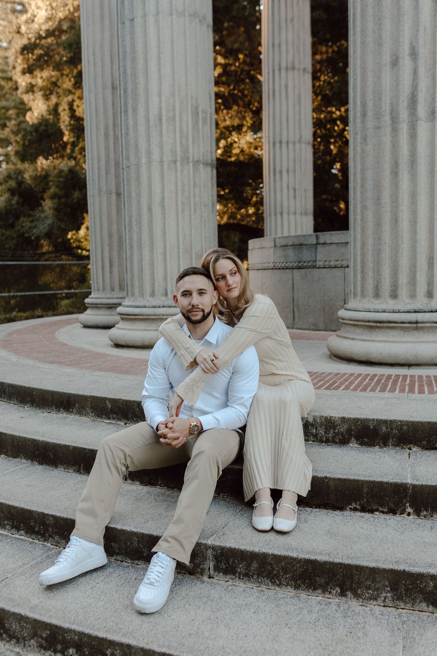 A couple in neutral colored clothing sits together on concrete steps with classic white columns in the background.