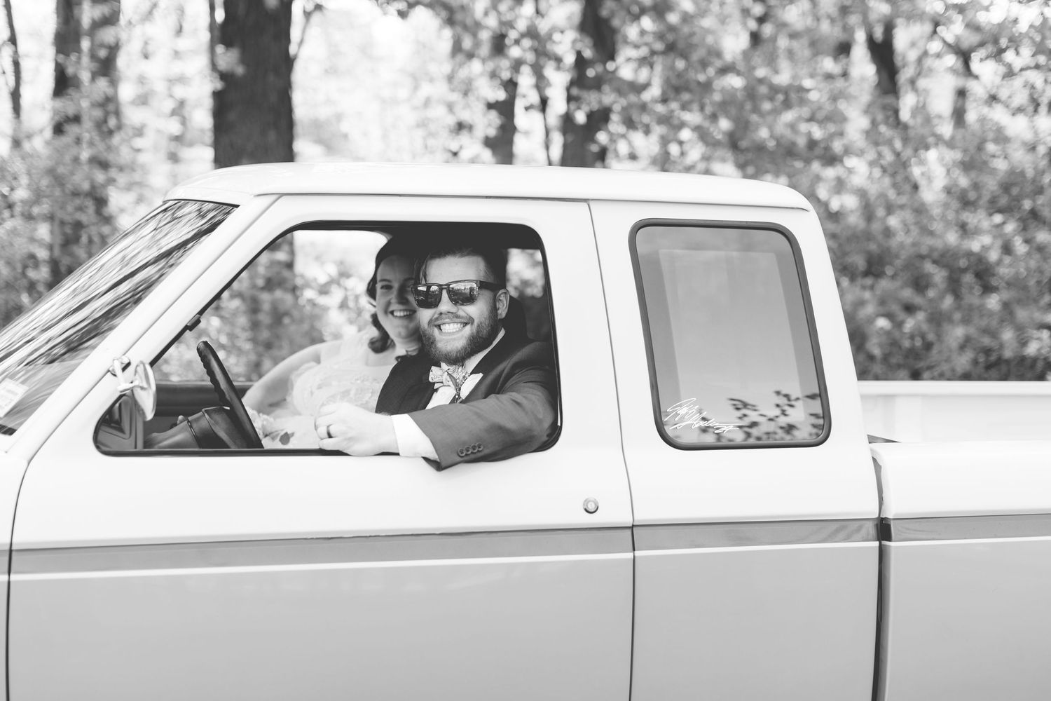 A person in sunglasses smiles while sitting in an old pickup truck in a black and white photograph.