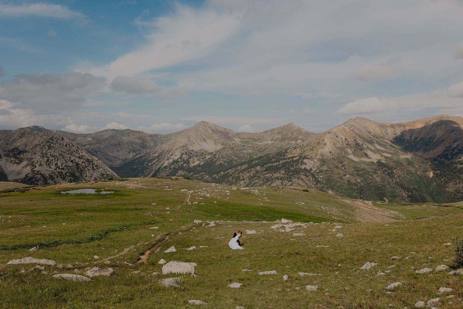 A winding mountain road cuts through green alpine meadows with rocky peaks and blue skies in the background.