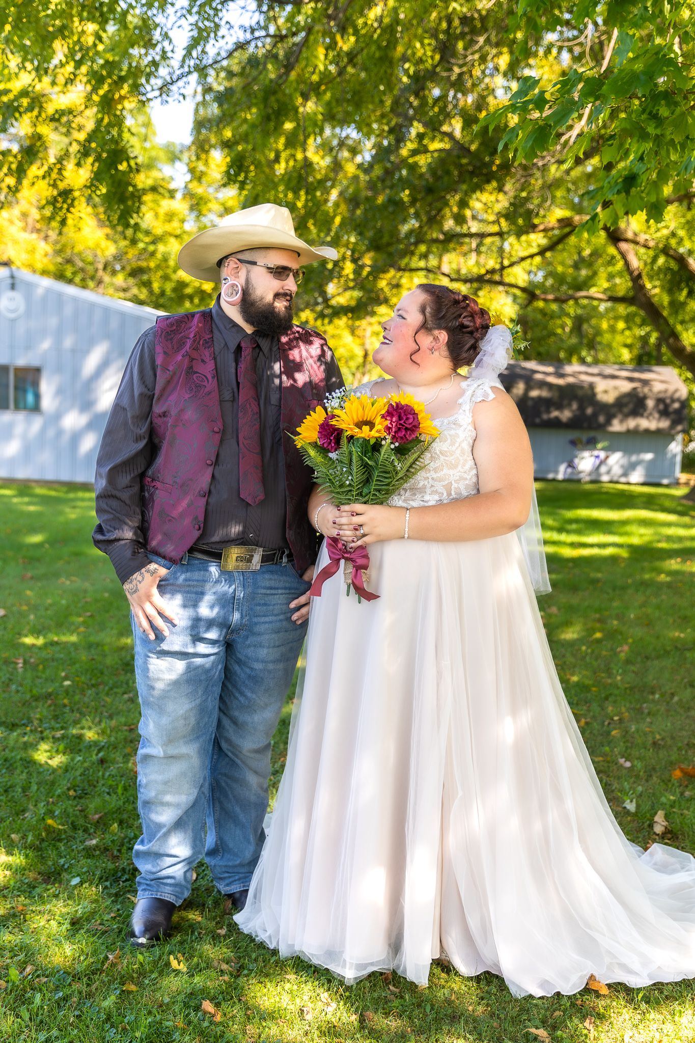 Wedding couple in rustic attire share a joyful moment with bright sunflowers on scenic grounds.