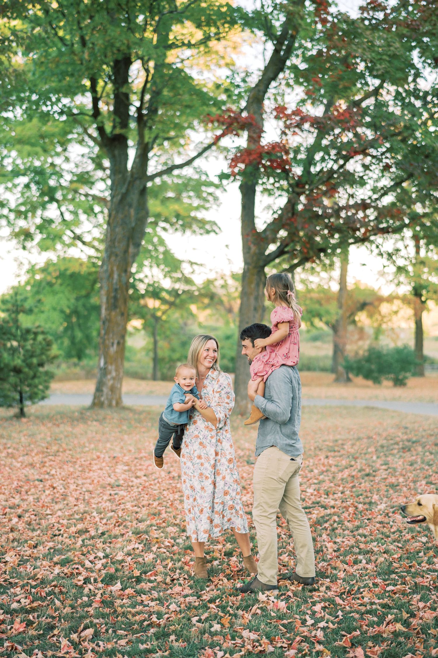 A happy young couple with children stand together in a forest setting filled with colorful autumn leaves.
