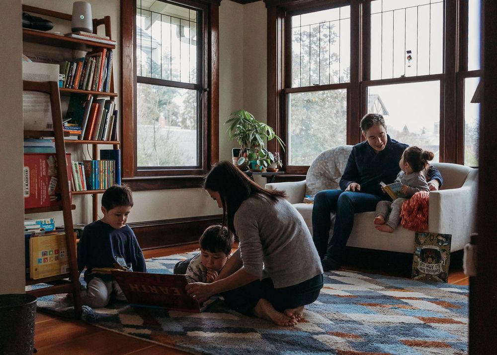 People gathered in a cozy living room with bookshelves and windows, spending time together on a comfortable carpet and couch.