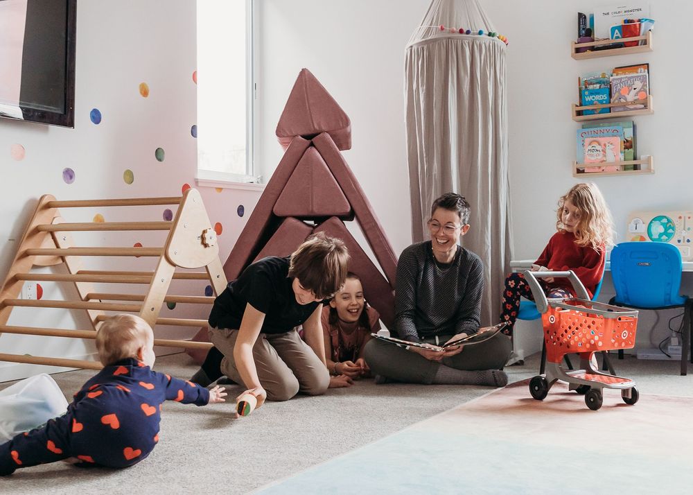 Kids play in a cozy playroom with wooden climbing triangle, tent and toys while adults sit together on the floor.