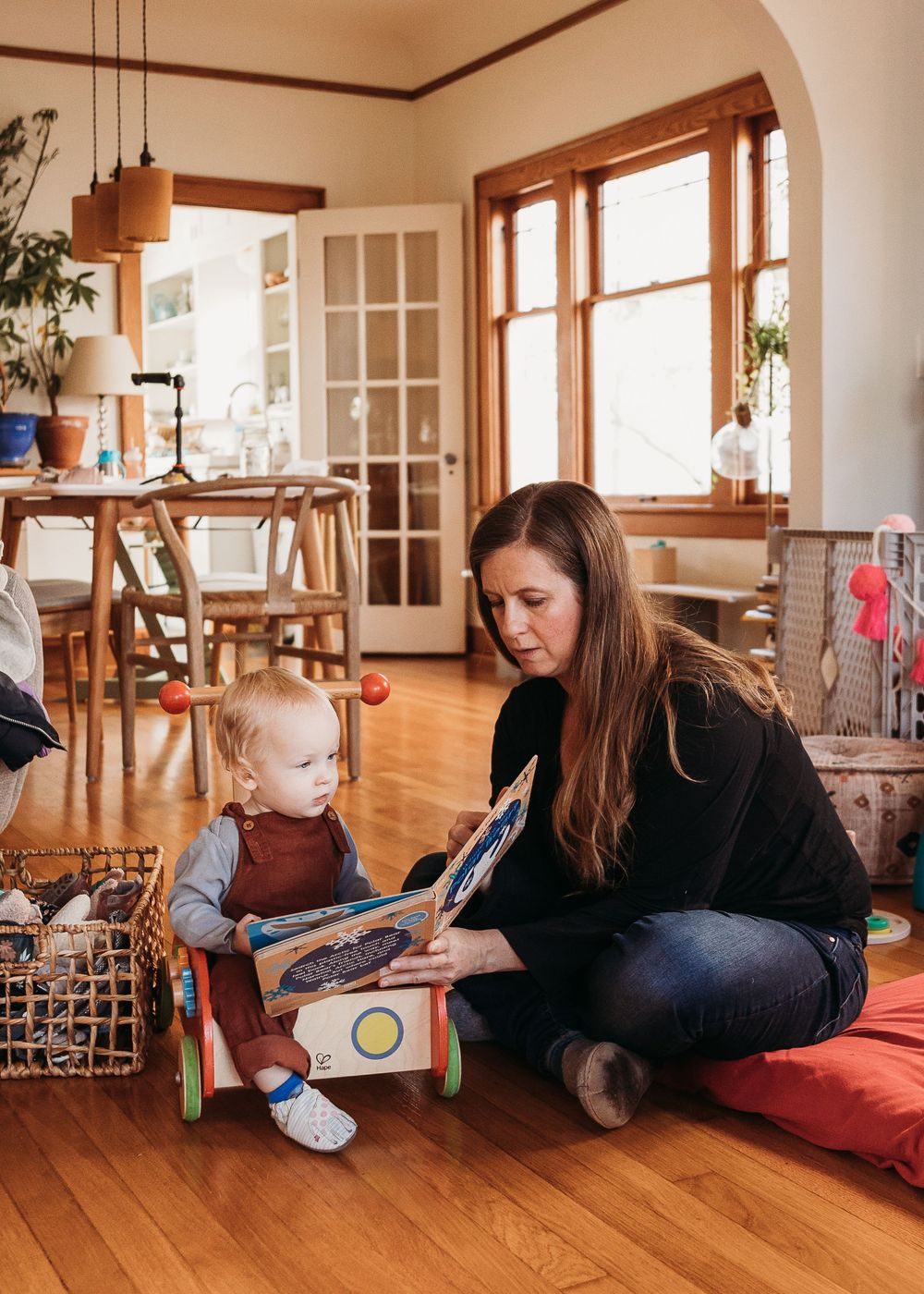 Adult and young child sit together on hardwood floor reading a book in cozy dining room with natural light.