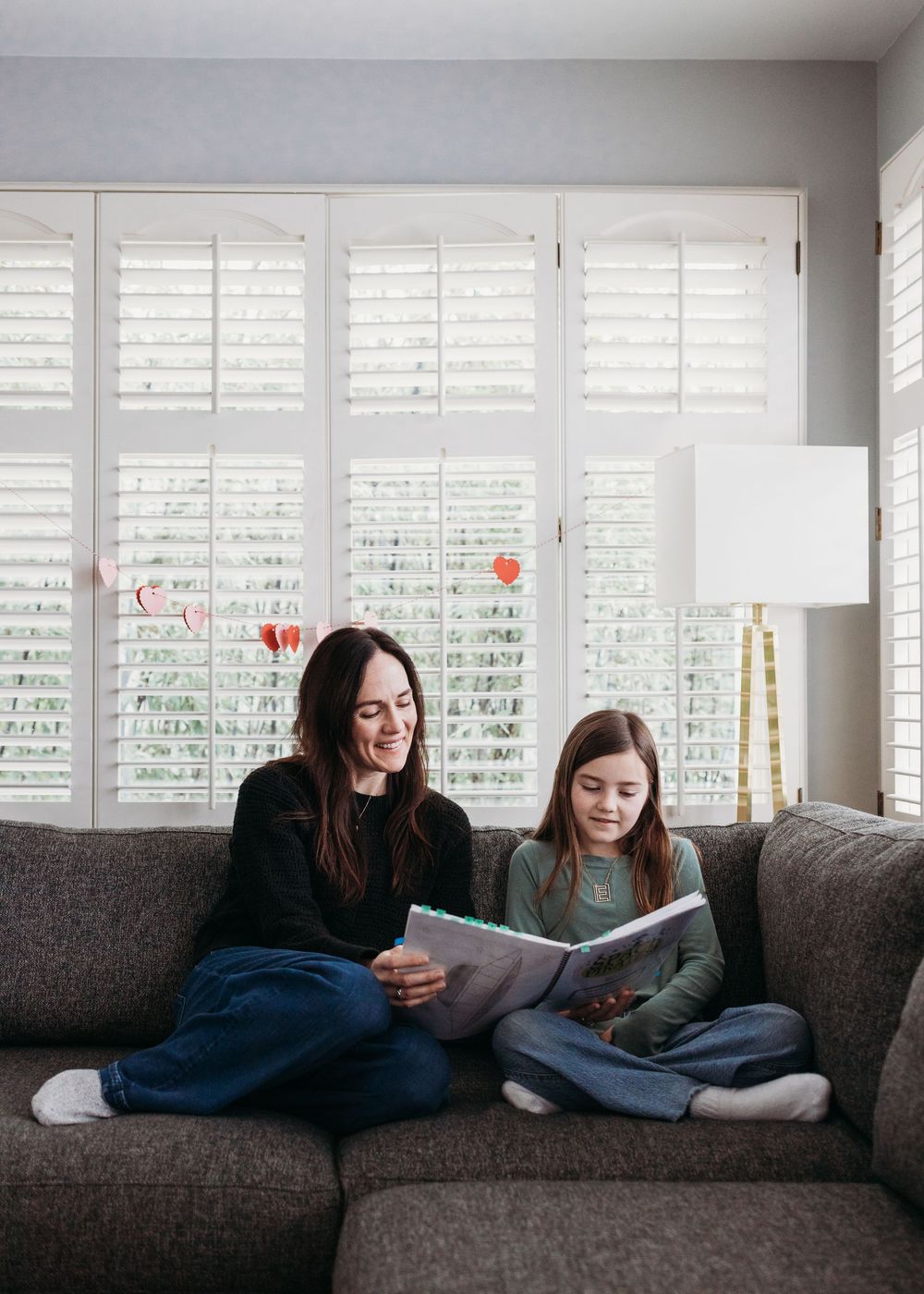 Two readers share a book together while relaxing on a gray couch near white shuttered windows.