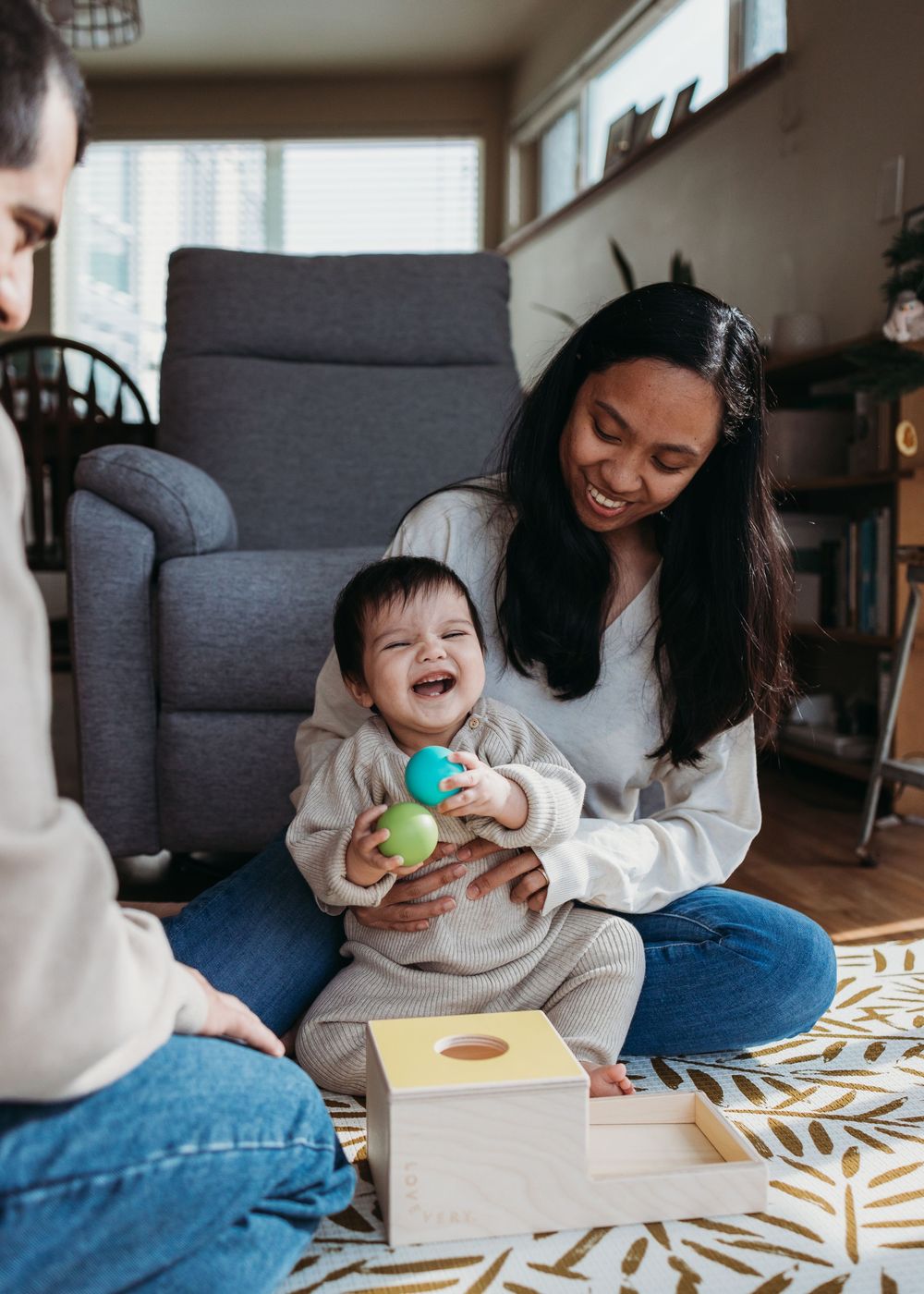 Family plays together with a wooden box toy on a patterned rug in a cozy living room setting.