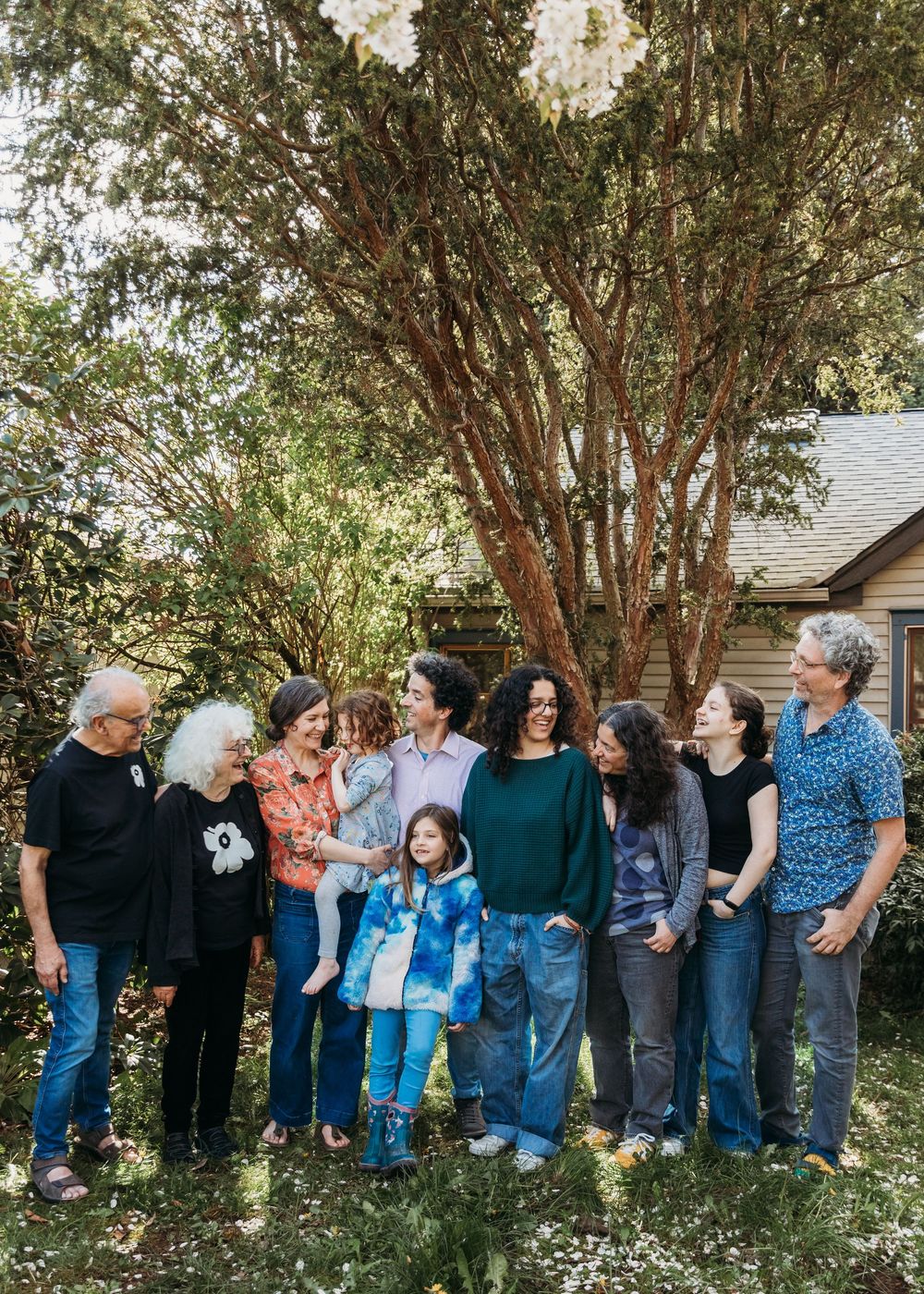 A multi-generational group gathers for a casual outdoor photo beneath a large tree on a sunny day.