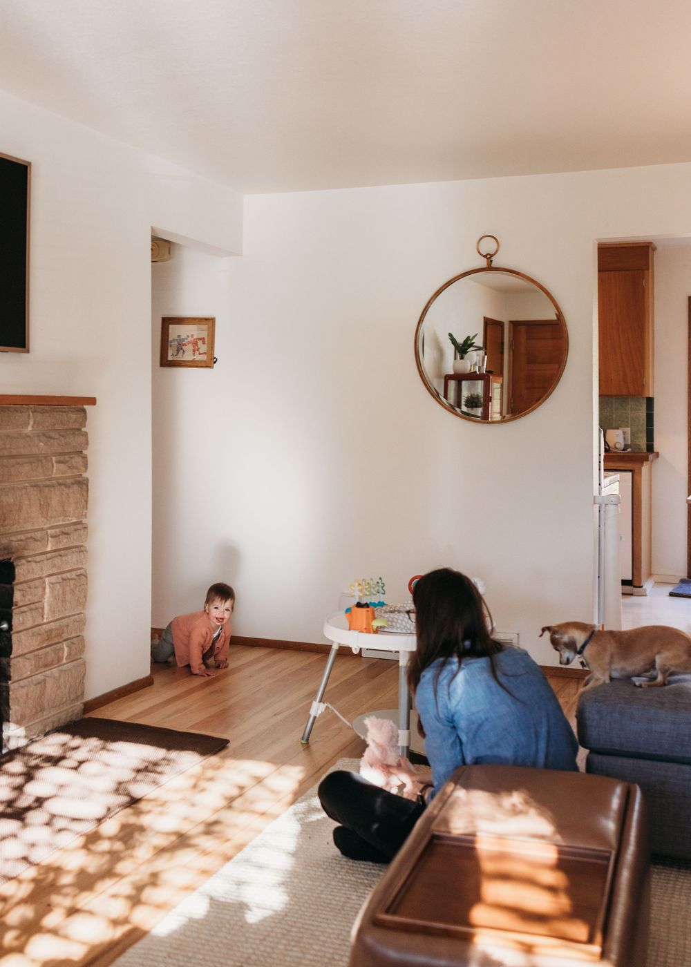 A cozy living room interior with dappled sunlight, round gold mirror, and people relaxing on furniture.