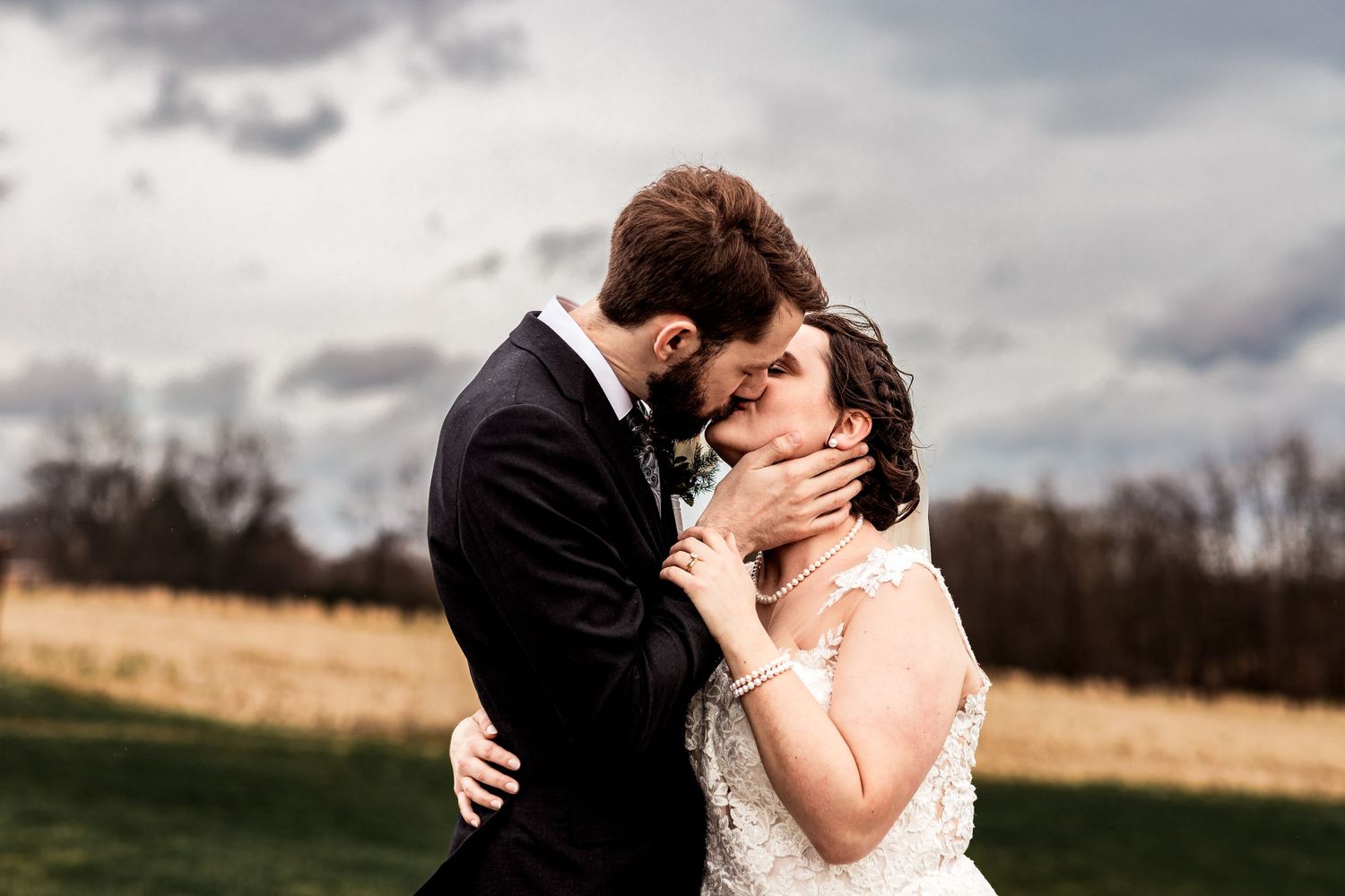 Romantic outdoor wedding photo of bride and groom kissing under cloudy sky, bride in lace gown and groom in dark suit.