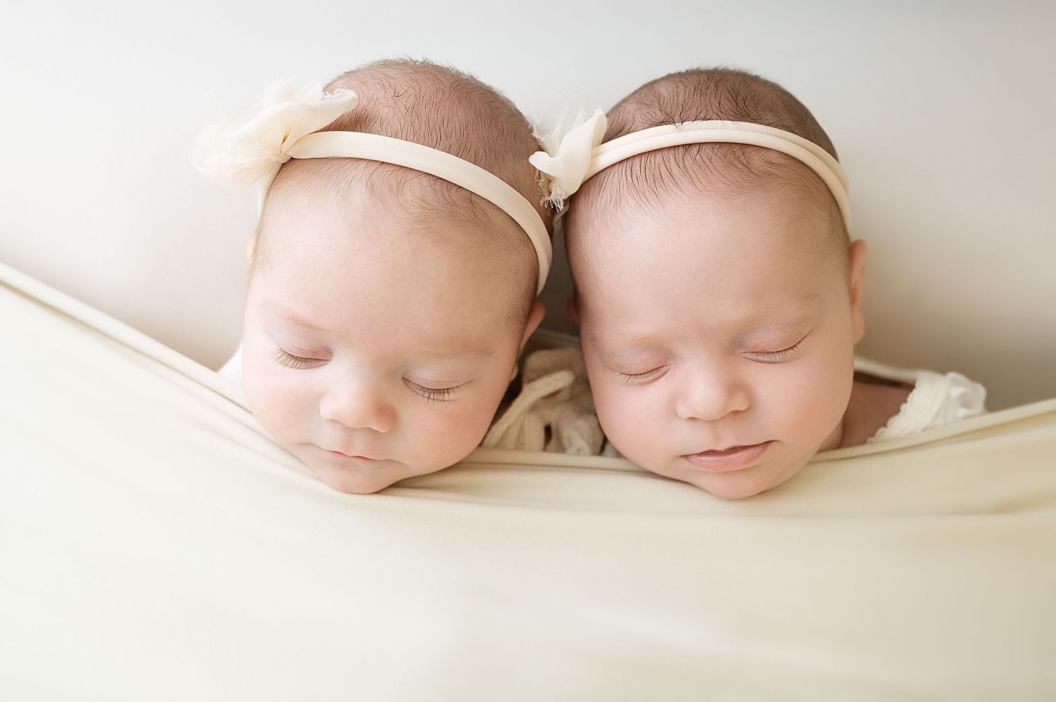 Two sleeping newborn babies swaddled in white blankets on a soft cream background.
