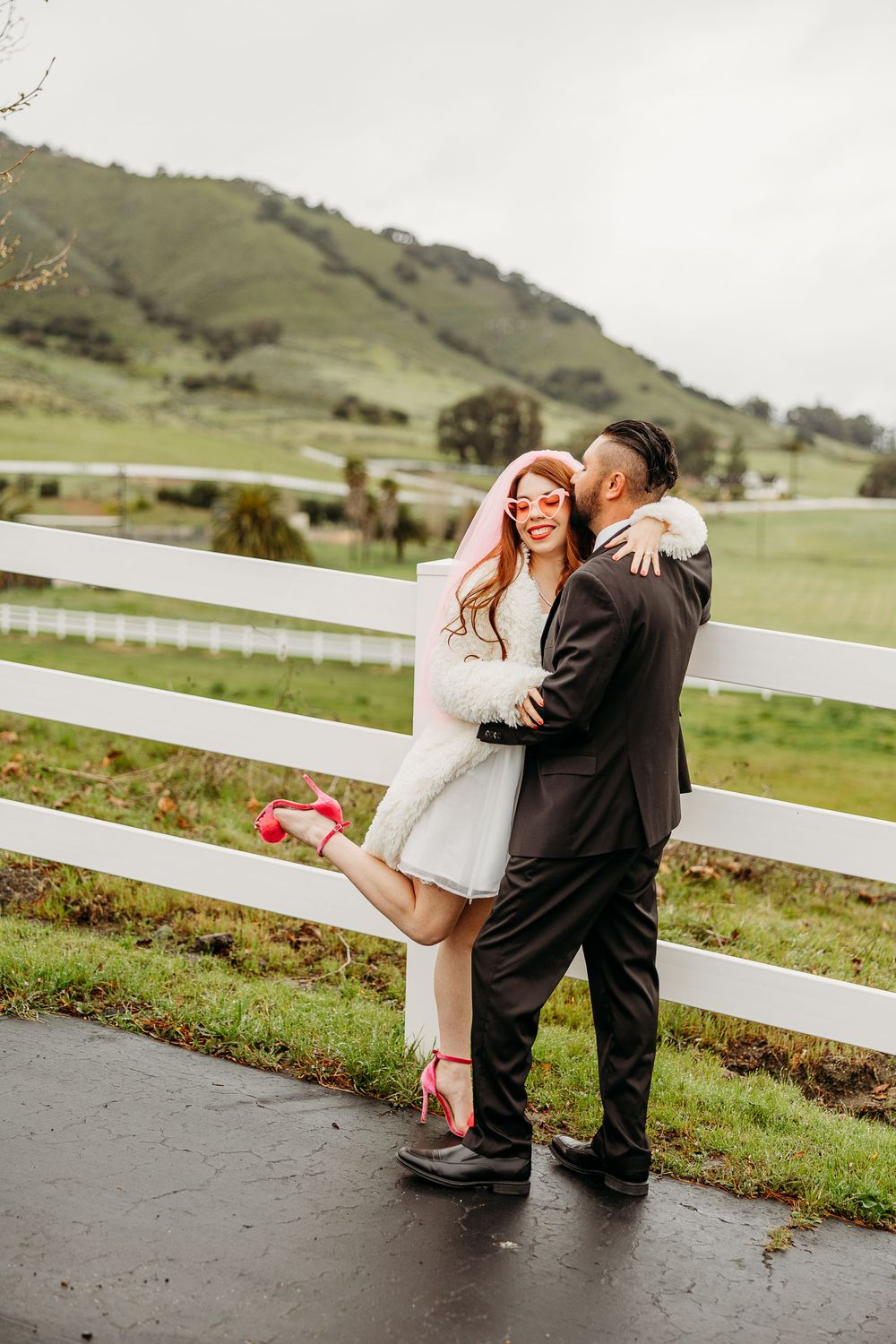 A couple shares a romantic moment by a white fence on a rainy day with green rolling hills in the background.