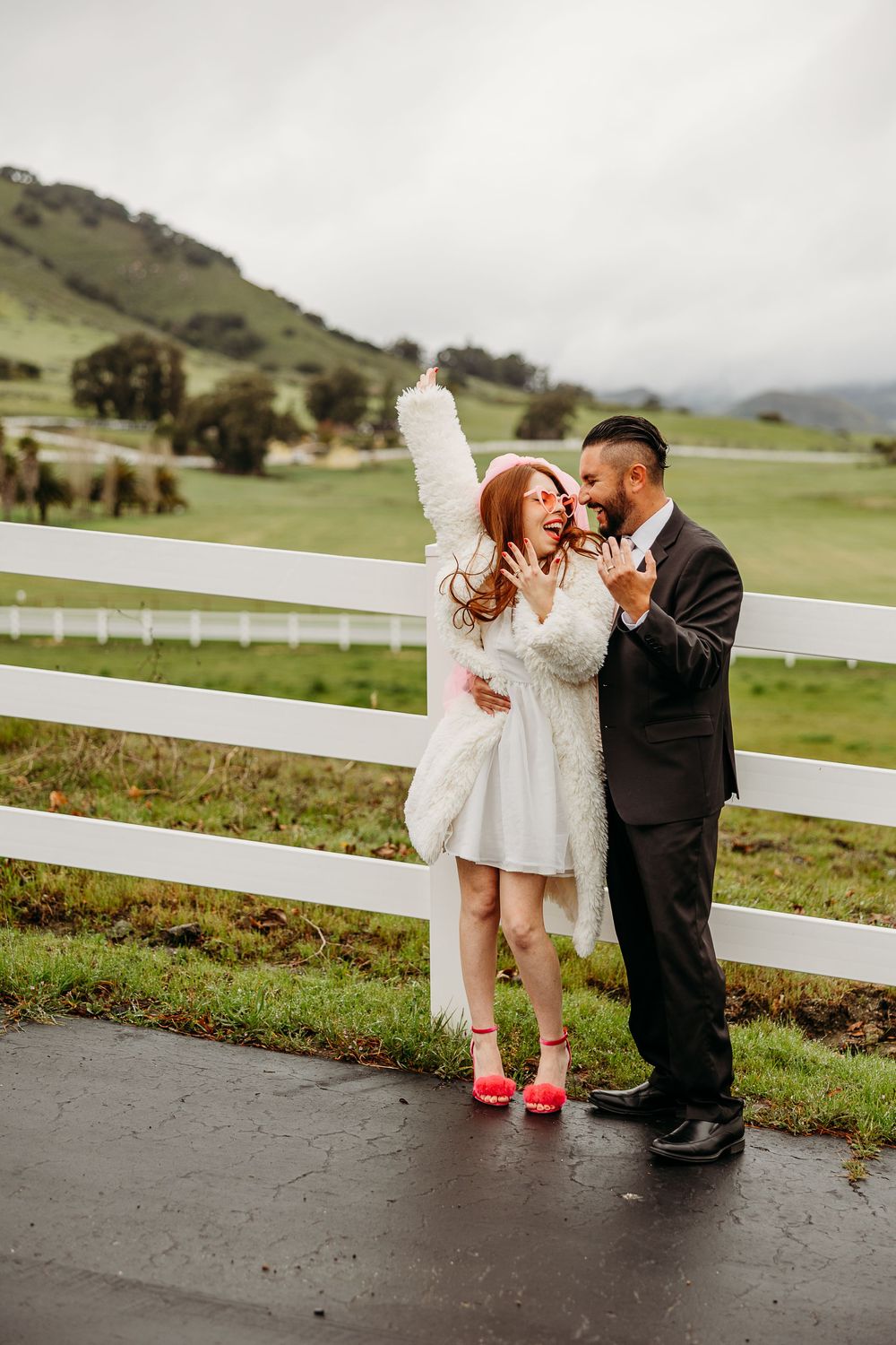 Children in formal wear play and interact together on a concrete walkway near a white fence with rolling hills in background.