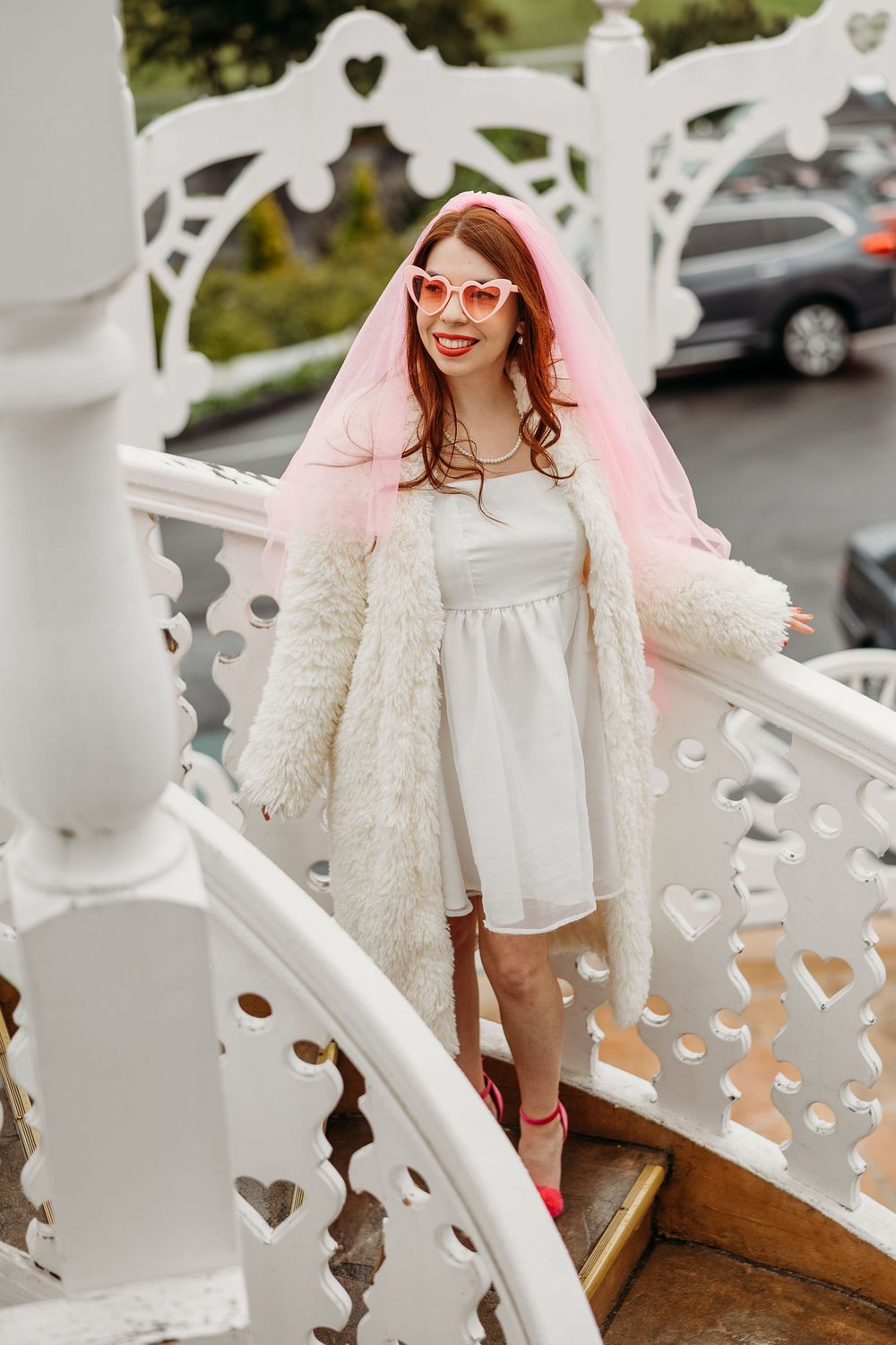 Person wearing a white dress, pink veil, and retro glasses stands on ornate white staircase during outdoor photo session.