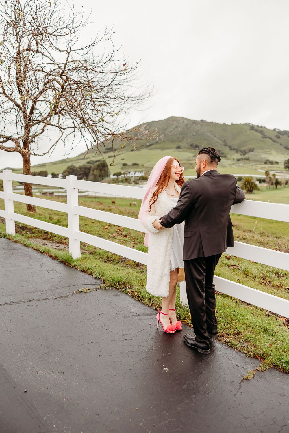 A couple stands embracing on a rainy path by a white fence with rolling green hills in the background.