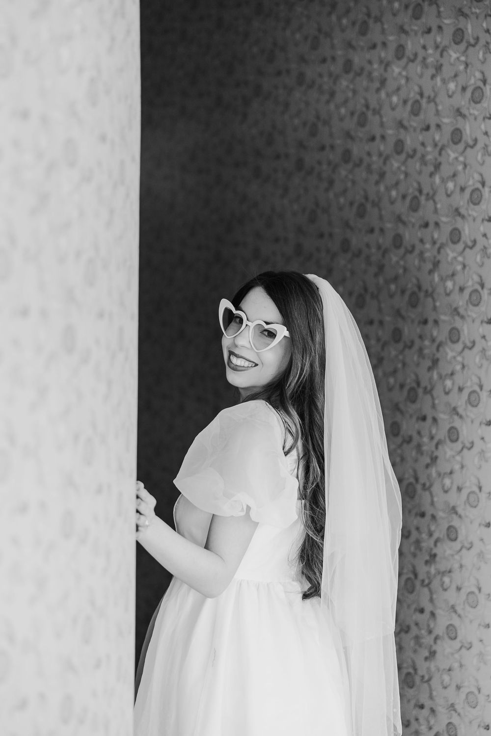 Black and white photo of a bride in glasses and wedding dress with tulle veil looking back over her shoulder.