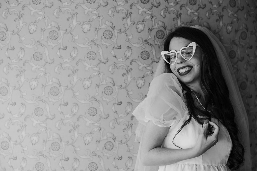Black and white portrait of a laughing person with glasses wearing a light colored dress against a patterned wallpaper.