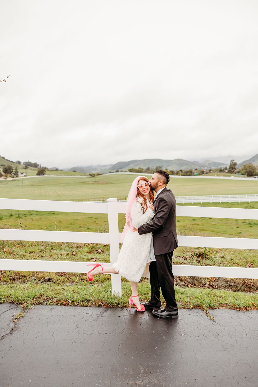 A romantic moment shared between a couple by a white fence on a rolling countryside farm with mountains in the background.