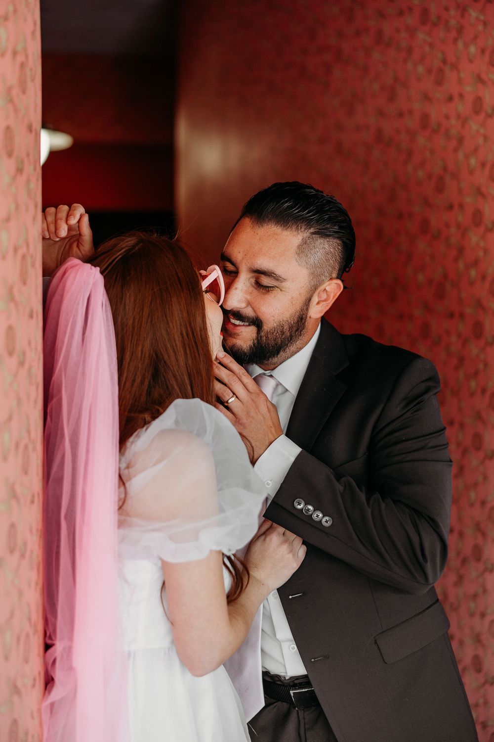 A romantic couple embraces against a deep red textured wall as they share an intimate moment together.