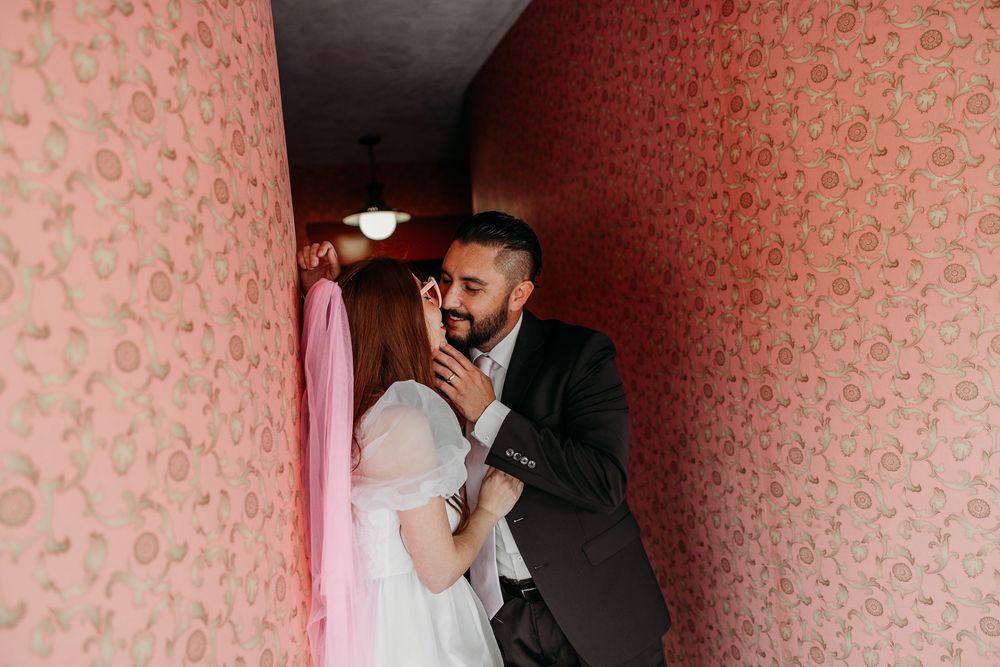 A couple shares an intimate moment in a hallway with pink floral wallpaper and dim lighting.