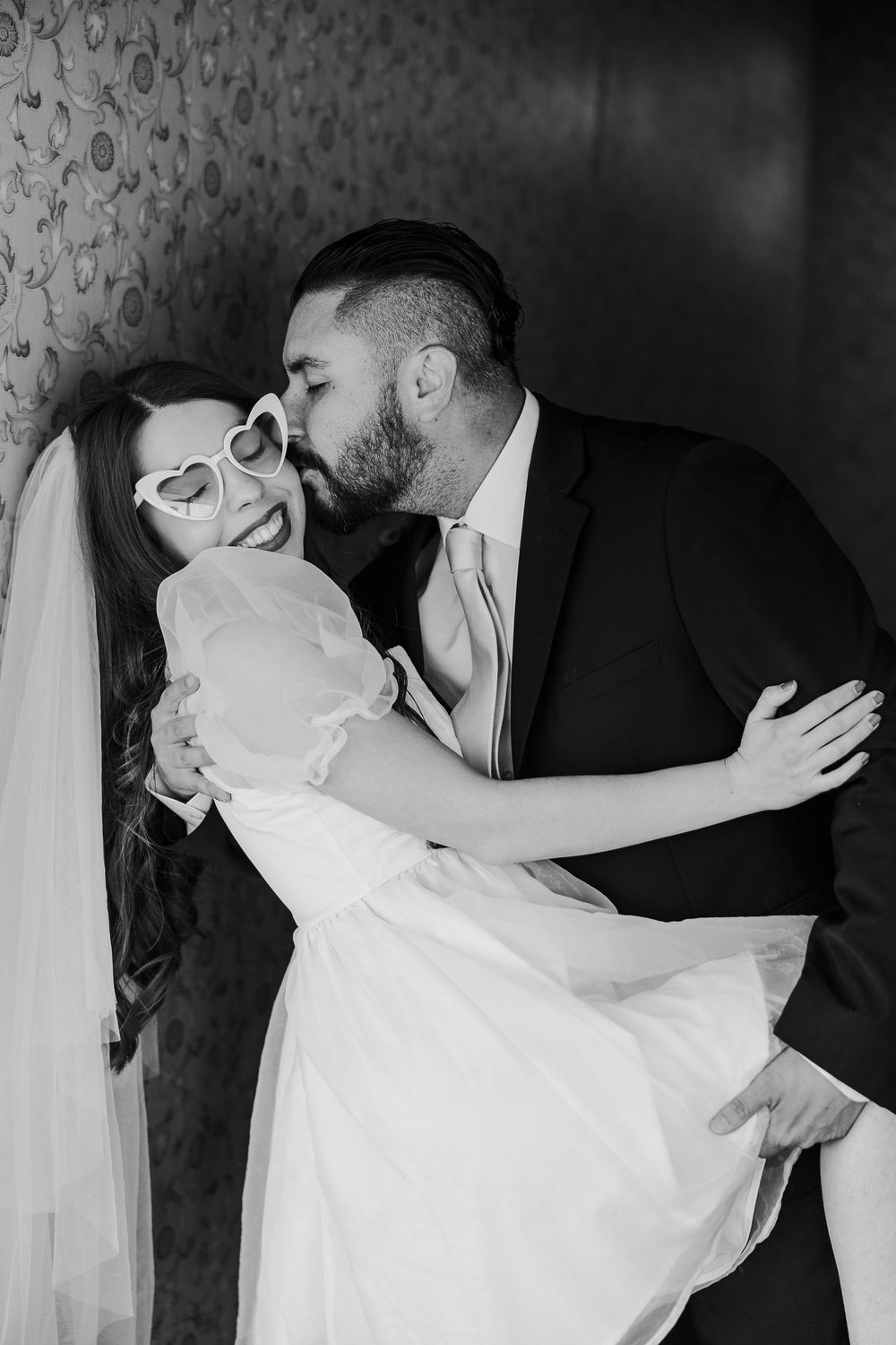 A romantic black and white wedding photo showing a couple in a dip pose against a patterned wallpaper background.