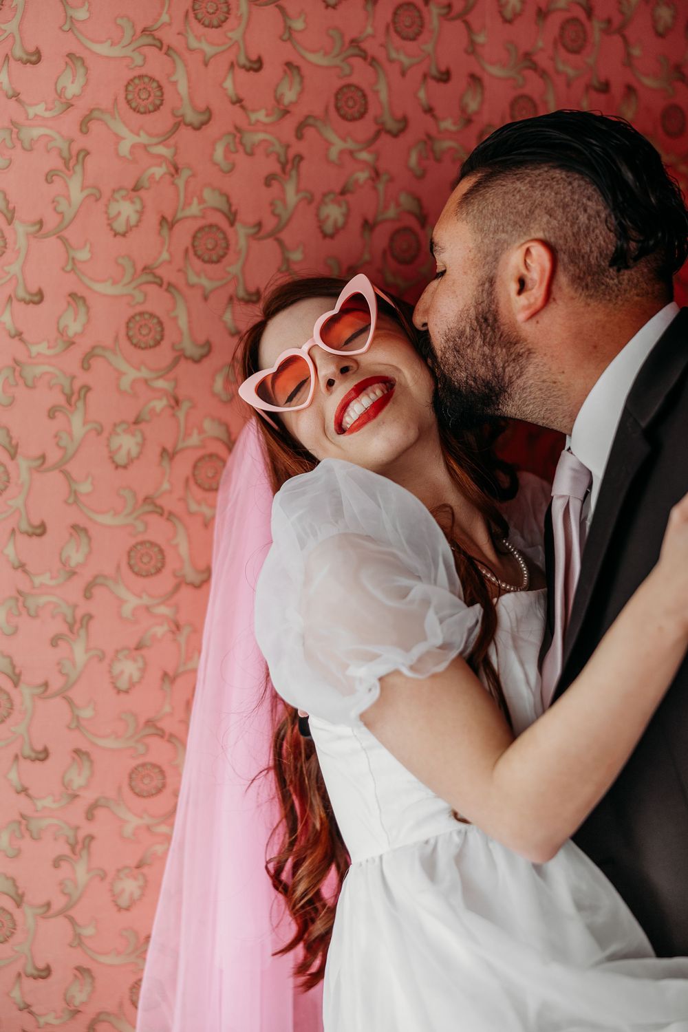 A romantic bride in heart-shaped sunglasses and pink hair laughs joyfully against a floral wallpaper backdrop.