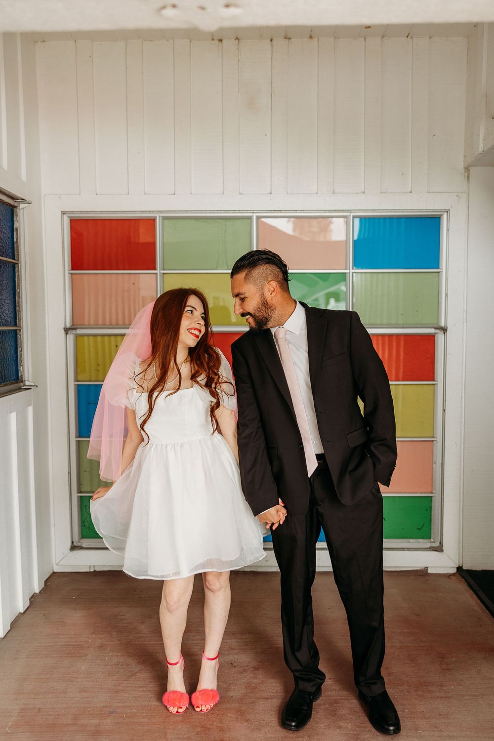 Couple in formal attire laughing together against a colorful paneled wall with pink and blue squares.